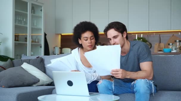Knappe Afro-Amerikaanse vrouw en knappe blanke man zittend op de bank in de woonkamer, met papieren bladen vol documenten en in vergelijking met de inhoud op het beeldscherm, tekst typen — Stockvideo