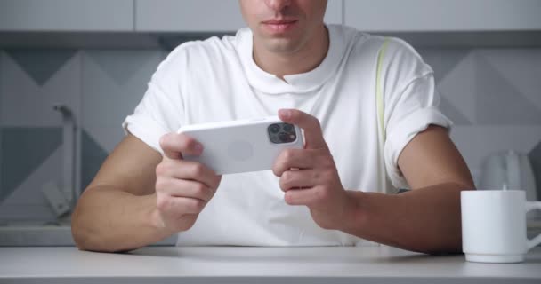 Close-up of unrecognizable young man playing on his smartphone exciting mobile application winning a racing game clenching fist rejoicing while sitting at kitchen table at home. — Stock Video