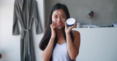 Nemlendirici yüz kremi olan güzel bir kadın yüzüne nemlendirici sürmüş bir kap tutuyor ve kurnazca gülümsüyor ve banyodaki bornozun arka planında asılı duran kameraya bakıyor.
