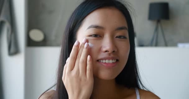 Gros plan d'une femme asiatique séduisante avec une peau saine, de beaux cheveux longs, appliquant une crème hydratante quotidienne sur son visage, effectuant un massage facial pour lisser pendant sa routine quotidienne du matin dans la salle de bain. — Video