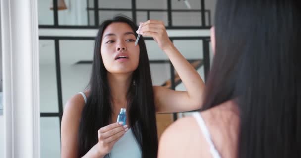 Çekici Asyalı kadın elinde anti-yaşlanma serumu ya da masaj yağı olan bir kap tutuyor, yüzüne damlalar damlatıyor, banyodaki ayna yansımasına bakarak masaj yapıyor. — Stok video