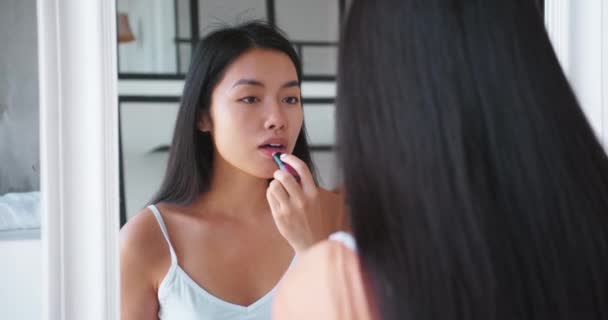 Retrato de una hermosa morena de pelo largo alegre asiática que aplica bálsamo higiénico en los labios, sonriendo, admirándose mirando su reflejo en el espejo del baño. Concepto de belleza y maquillaje — Vídeos de Stock