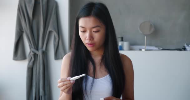 妊娠検査のストリップを保持している楽しい若いアジアの可愛い女性は、結果がバスルームに立って、素晴らしい結果の後に喜びを待っています。排卵、不妊、婦人科医療の概念 — ストック動画