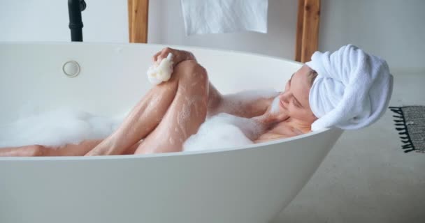 浴槽の上に寝そべっている頭にバスタオルをつけた魅力的な女性、白い家庭用バスルームで泡風呂をしながら洗濯やマッサージのための洗濯布を使用して、スパで休日を楽しんで — ストック動画
