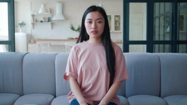Portret van een zelfverzekerde jonge Aziatische vrouw die thuis op de bank zit. Charmante jonge Aziatische etniciteit vrouw kijken vol vertrouwen in de camera, zitten op de bank thuis — Stockvideo