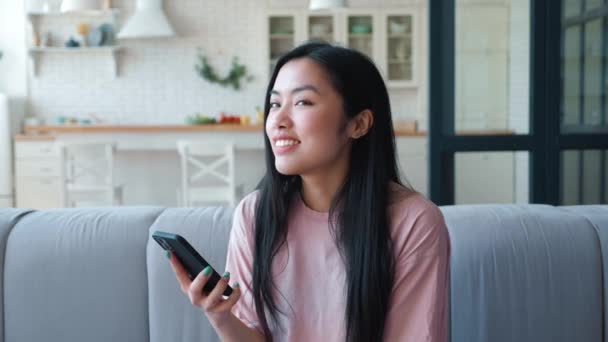 Sjarmerende og mystisk tenkende ung asiatisk kvinne som ser seg rundt mens hun sitter på sofaen og skriver meldinger fra smarttelefonen. Vakker kvinne som tekster med mobiltelefon – stockvideo