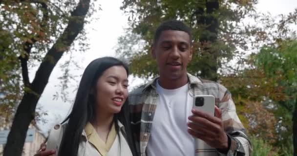Hermosa pareja heterosexual amorosa, dos amigos caminando por la calle cerca el uno del otro y mirando la pantalla de un teléfono inteligente en las manos de un chico africano sonriente feliz. Relaciones humanas — Vídeos de Stock