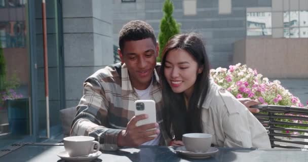 Young multiracial loving couple sitting together in the outdoor cafe on beautiful sunny day at romantic date , taking a self portrait and posting image on social media. — Stock Video