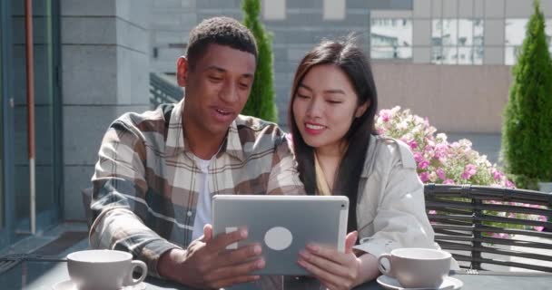 Beautiful loving couple, two close friends sitting in the outdoor cafe on warm sunny day at date, talking looking at digital tablet in the hands of handsome guy, swiping, surfing on web pages — Stock Video