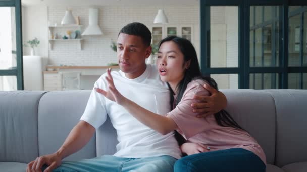 Happy family interracial young heterosexual couple, African guy and Asian girl sitting close to each other, embracing ang having fun discussion, pointing to the side and enjoying time together at home — Stock Video