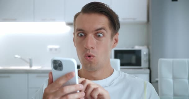 Verrast jonge man winnaar hold smartphone lees goed nieuws verbaasd door mobiele online bet game win thuis. Gelukkig mannelijk kijken naar mobiele telefoon scherm dolblij met de overwinning succes tijdens het zitten in de keuken — Stockvideo