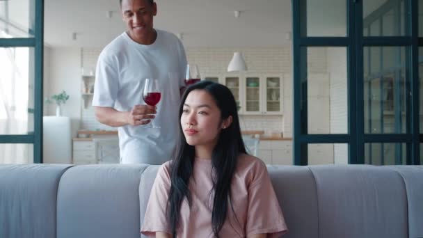 Veselý smíšený závod Afroameričan macho chlap se připlíží zezadu a přinést sklenici s červeným vínem na jeho přítelkyně asijského původu, cinknutí sklenice s červeným vínem na rande, těší trávit čas spolu — Stock video