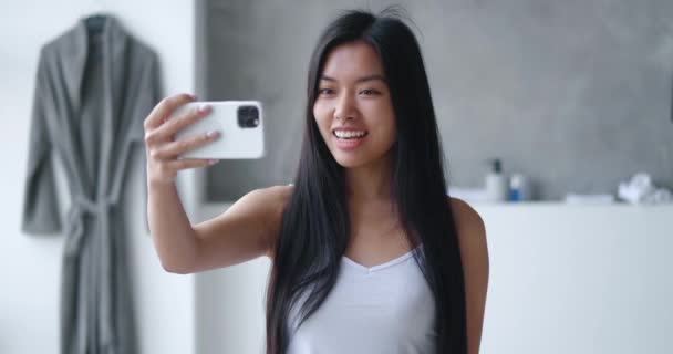 Jeune femme asiatique faisant appel vidéo à l'aide d'un smartphone dans la baignoire. Millennial fille dans le bain avec téléphone portable ayant webcam conversation vidéo profiter des soins corporels quotidiens dans la salle de bain — Video