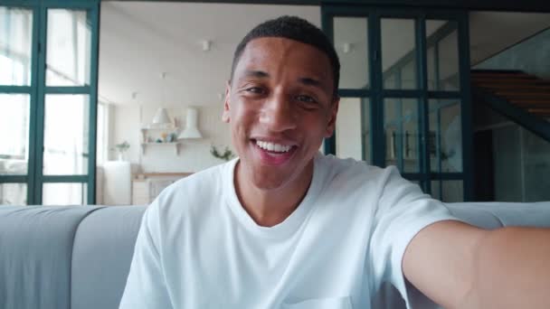 Χαμογελώντας Αφροαμερικανός που κάνει selfie ή βιντεοκλήση ενώ κάθεται στο σπίτι στον καναπέ. Πορτρέτο του νεαρού άνδρα blogger κάνει μια συνομιλία βίντεο από το διαμέρισμά του, ενώ κάθεται στον καναπέ — Αρχείο Βίντεο