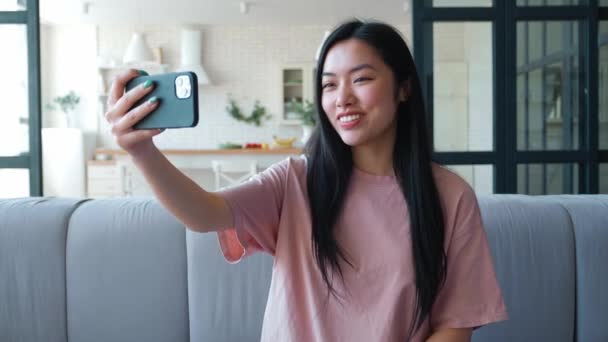 Mosolygó gyönyörű ázsiai nő kezében okostelefon és így szelfi vagy videó hívást, miközben otthon ül a kanapén. Fiatal nő blogger csinál video chat mobiltelefon a lakásából — Stock videók