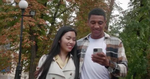 Encantadora pareja joven y cariñosa, dos amigos caminando por la calle cerca uno del otro y mirando la pantalla de un teléfono inteligente en manos de un chico africano feliz y sonriente. Conceptos de relaciones humanas — Vídeos de Stock