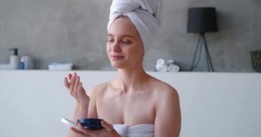 Banyoda beyaz banyo havlusuna sarılı güzel bir kadının, güzellik ürünleriyle dolu bir kabı açarken, nemlendirici krem sürerken ve vücuduna nemlendirici sürerken, rahatlatıcı masaj yaparken.