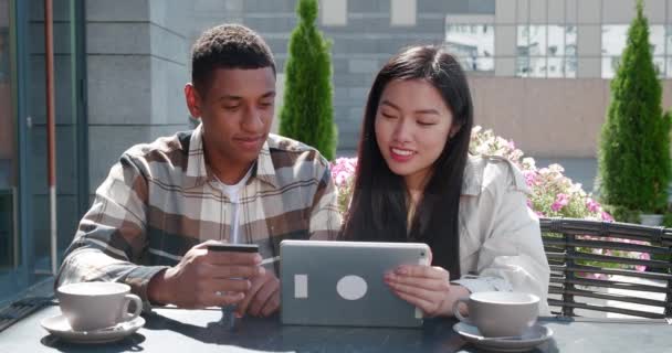 Szczęśliwa para Afrykańczyków trzymających kartę kredytową i wesołą Azjatkę z tabletem, siedzącą przy stole letniego tarasu przytulnej kawiarni, patrzącą na cyfrowy tablet i robiącą zakupy online — Wideo stockowe