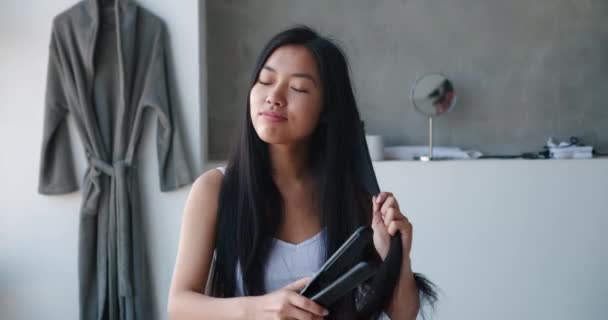 Düz saç. Güzel gülümseyen Asyalı kadın, sabah güzellik ve hijyen rutininden zevk alır, şarkı söyler ve dans ederken düz ütüyle sağlıklı uzun saçlar düzleştirir, ev tuvaletinde saç düzleştirici — Stok video