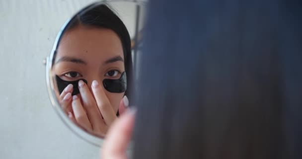 Regelbunden hudvård. Glada unga asiatiska vacker kvinna står i badrummet, tittar på hennes reflektion i en liten sminkspegel tillämpa svart utjämning och återfuktande kollagen fläckar under ögonen — Stockvideo