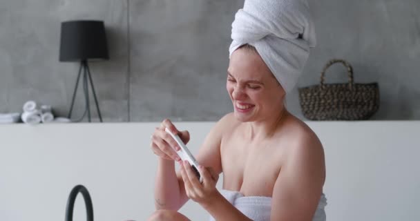 Joven mujer sonriente jugando aplicación de juego en el teléfono inteligente mientras está sentado en el baño disfrutando del día de belleza — Vídeos de Stock