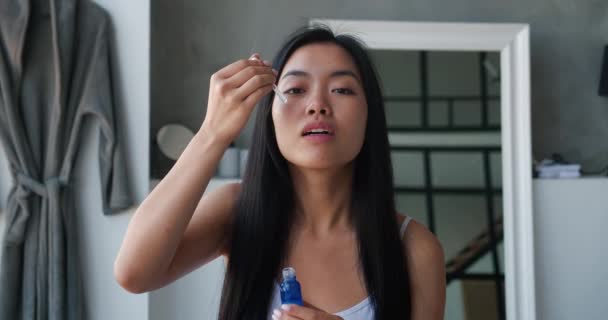 Hermosa mujer asiática sosteniendo un frasco con suero hidratante o aceite de masaje y aplicando unas gotas en su cara usando un gotero, realizando un masaje suavizante mirando su imagen en el espejo del baño — Vídeos de Stock