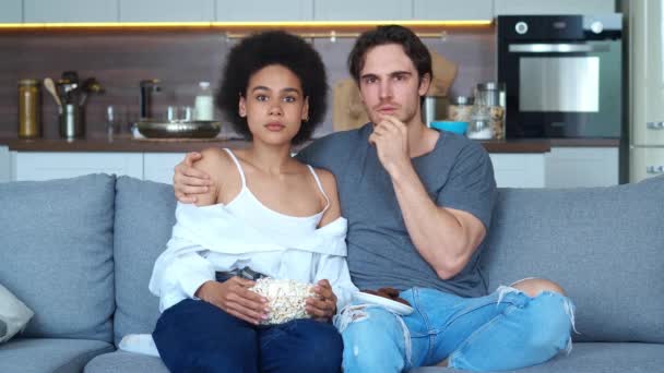Skockade par av kaukasisk man och afrikansk amerikansk ung kvinna tittar på skräckfilm film på eleganta lägenheter, sitter på soffan tillsammans med popcorn hemma — Stockvideo