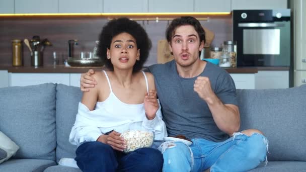 Podekscytowana młoda para, biały mężczyzna i afrykańska amerykańska kobieta oglądająca futbol w domu. Świętuj zwycięstwo, zwycięstwo drużyny telewizji bramkowej w meczu telewizyjnym siedzi na kanapie z popcornem — Wideo stockowe