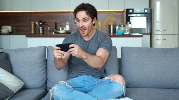 スマートフォンで遊ぶ若い男エキサイティングなモバイルアプリケーションは、自宅のリビングルームでソファに座っている間に喜びをレースゲームのクリーンアップを受賞. — ストック動画