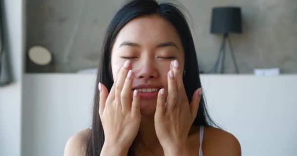 Primer plano de una encantadora mujer asiática que se aplica una crema hidratante nutritiva en la cara y realiza un masaje facial suavizante con los dedos y sonriendo mirando a la cámara. Tratamiento de higiene matutina — Vídeos de Stock