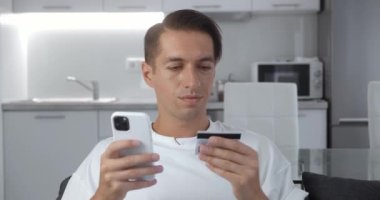 Çekici beyaz adam elinde kredi kartıyla evde akıllı telefon kullanarak kanepede oturuyor ve gülümsüyor. Finans, internet üzerinden alışveriş.