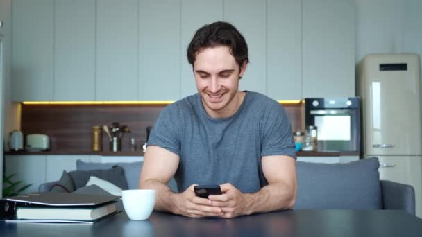 Guapo chico macho con un aspecto perfecto, peinado limpio, sosteniendo el teléfono inteligente, sonriendo mientras escribe mensaje de texto en la pantalla táctil, sentado en el sofá en su apartamento estudio. Hombre descansando en casa — Vídeo de stock