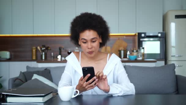 Młoda Afroamerykanka coquettishly wpisując tekst w smartfonie, patrząc obsesyjnie na bok, z kochającym spojrzeniem, nadal pisze wiadomość siedząc przy stole z uśmiechem na twarzy — Wideo stockowe