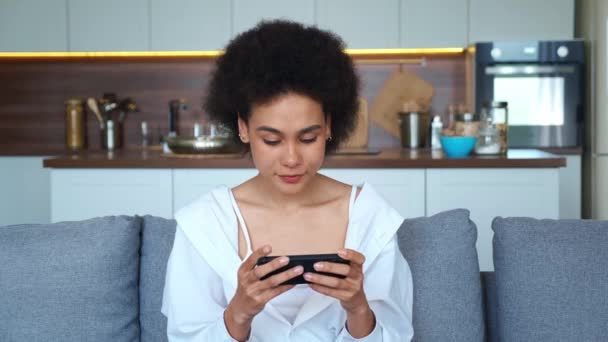 Jong afrikaanse amerikaanse vrouw spelen op haar smartphone spannende mobiele applicatie racespel terwijl zitten op de bank in de woonkamer thuis. — Stockvideo