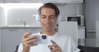 Evdeki koltukta oturmuş akıllı telefonuyla komik videolar izleyen kulaklıklı neşeli genç bir adam. Erkek cep telefonu kullanmaktan hoşlanıyor.