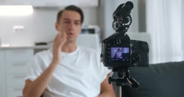 Blogueur millénaire ou vlogger homme filmant nouvelle vidéo vlog avec caméra professionnelle, monté sur trépied assis sur le canapé à la maison. — Video