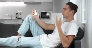 Akıllı telefonlu gülümseyen genç adam oturma odasında kanepede uzanırken video görüşmesi yapıyor. Evdeki kanepede dinlenen mutlu bir adamın portresi.