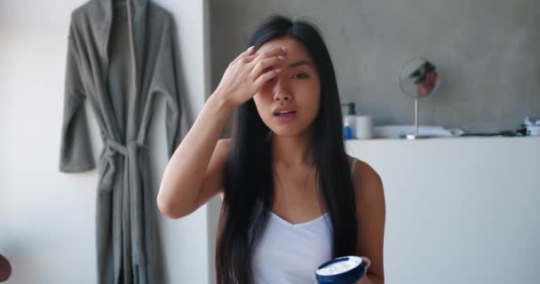 Fröhliche junge hübsche Asiatin, die sich Feuchtigkeitscreme ins Gesicht sprüht, den Container mit dem Schönheitsprodukt jubelnd in die Kamera blickt und im Badezimmer mit hängendem grauen Bademantel tanzt — Stockvideo