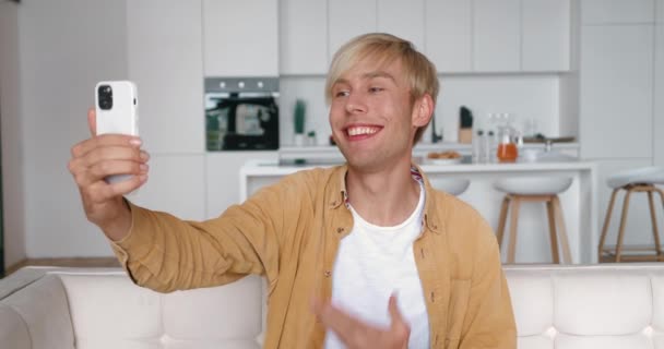 Hombre joven hablando con la cámara del teléfono inteligente al tener videocall en el interior. Retrato de un hombre sosteniendo el teléfono inteligente y videochat y sonriendo mientras está sentado en el sofá en casa — Vídeos de Stock