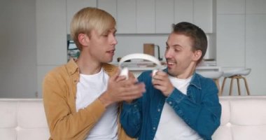 Erkek eşcinsel çift evde vakit geçiriyor. İki LGBTQ adamı bir koltukta dinleniyor ve Smartphone ve oturma odasındaki kulaklıkla müzik dinliyor.
