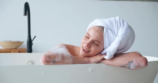 Belle femme blonde aux yeux fermés de plaisir, enveloppant sa tête dans une serviette de bain, couchée dans la baignoire et profitant d'une baignoire avec mousse au spa à la maison. Soins du corps et de la peau, concept d'hygiène et de relaxation. — Video