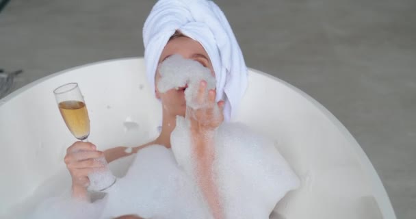 Κοντινό πλάνο πορτρέτο της ελκυστικής σέξι γυναίκας ξαπλωμένη γυμνή στην μπανιέρα γεμάτη αφρό στο μοντέρνο λευκό μπάνιο, απολαμβάνοντας όμορφη ημέρα ομορφιάς δοκιμάζοντας νόστιμη σαμπάνια από το κύπελλο και τη φροντίδα του δέρματος της. — Αρχείο Βίντεο