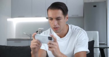 Akıllı telefonuyla oynayan genç adam heyecan verici mobil uygulaması evdeki oturma odasında kanepede otururken yumruğunu sıkarak bir yarış oyunu kazanıyor..