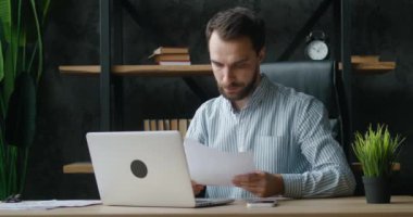 Modern işyerinde kağıt belgeler ve dizüstü bilgisayarla çalışan odaklanmış bir iş adamı. Bilgisayarı kullanan erkek girişimci, modern ofiste grafik iş süreci ile çalışıyor