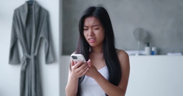 Zszokowana Azjatka reagująca na złą wiadomość na smartfonie stojącym w wannie. Nieprzyjemnie zaskoczona dziewczyna zdenerwowana utratą telefonu komórkowego na tle domowej łazienki — Wideo stockowe