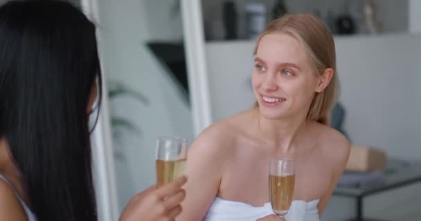 Close-up of two delighted beautiful multi-ethnic women chilling together at hen-party in home bathroom, sitting on bathtub and holding flute of champagne. Focus on European blonde smiling cute woman — Stock Video