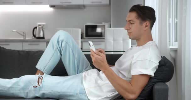携帯電話で現代のアパートのテキストメッセージでスマートフォンの笑顔を使用してソファの上に休んで陽気な若い男の閉じる,スクロールタップ技術 — ストック動画
