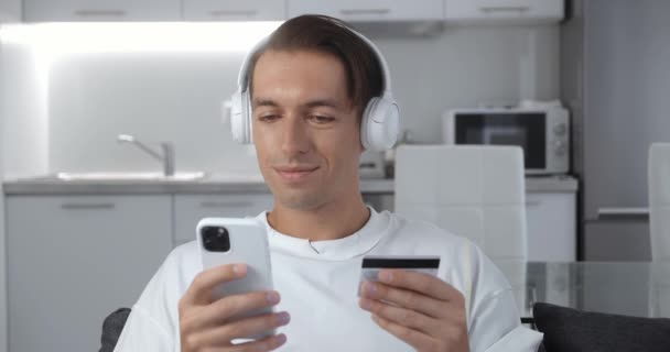 Młody człowiek w słuchawkach słucha muzyki i dokonuje płatności online trzymając kartę kredytową za pomocą smartfona w domu, siedzi na kanapie i uśmiecha się. Finanse, zakupy przez Internet. — Wideo stockowe