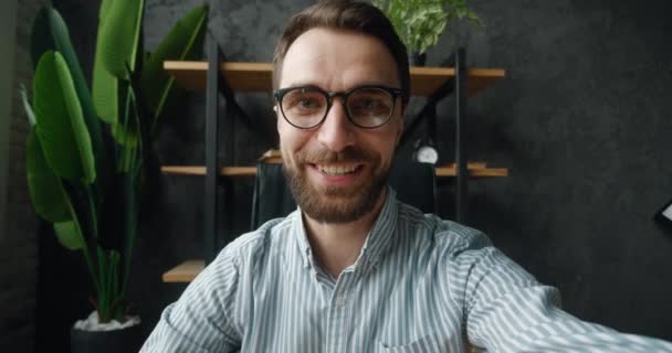 POV του όμορφος επιχειρηματίας κάνει selfie βιντεοκλήση, ενώ κάθεται στο σύγχρονο γραφείο. Πορτρέτο ενός επιχειρηματία blogger σε γυαλιά ηλίου κρατώντας smartphone, γυρίσματα blog βίντεο σε εσωτερικούς χώρους στο γραφείο στο σπίτι — Αρχείο Βίντεο