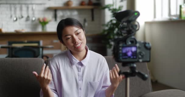 Young and attractive asian woman recording video blog with professional video camera mounted on tripod. Happy female blogger sitting on sofa at home and talking to camera while recording vlog video — Stock Video